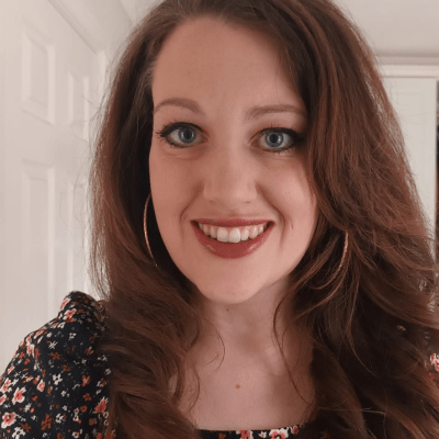 A smiling woman with long, wavy brown hair dressed in a floral top, standing indoors in front of a white wall.